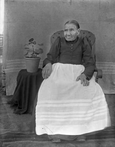 Portrait of Mrs. William Krueger, sitting, with a potted geranium plant placed on a table behind her. A blanket is hanging behind her as a backdrop.