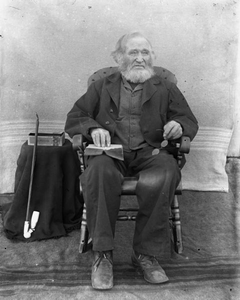 Portrait of William Krueger holding a book in his lap and holding a pair of eyeglasses. Behind him, a long pipe is resting against a table on which a box is sitting, probably of tobacco. A blanket is hanging behind him as a backdrop.