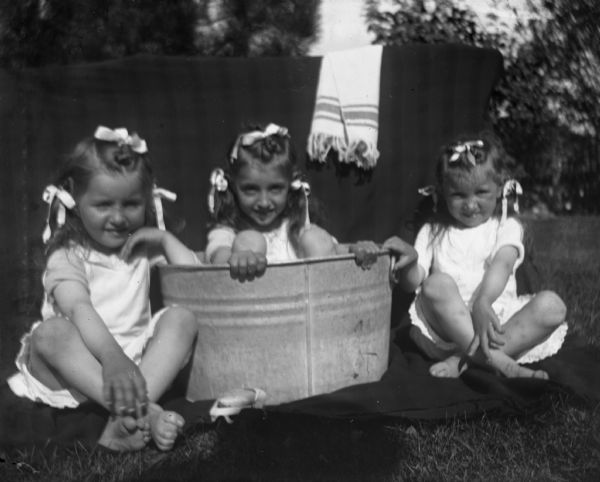 Jose and Esther Bigalk sitting with Emma Gercke outdoors during the summer months with bows in their hair. One girl is sitting in a wash basin and the others are sitting beside her.