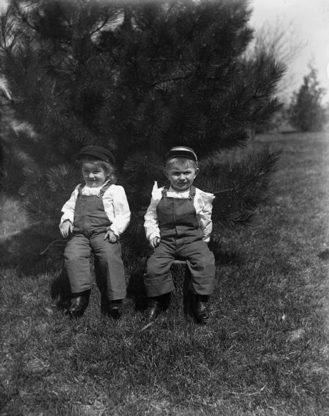 Portrait of Jennie and Edgar Krueger, sitting on logs, wearing overalls and hats.