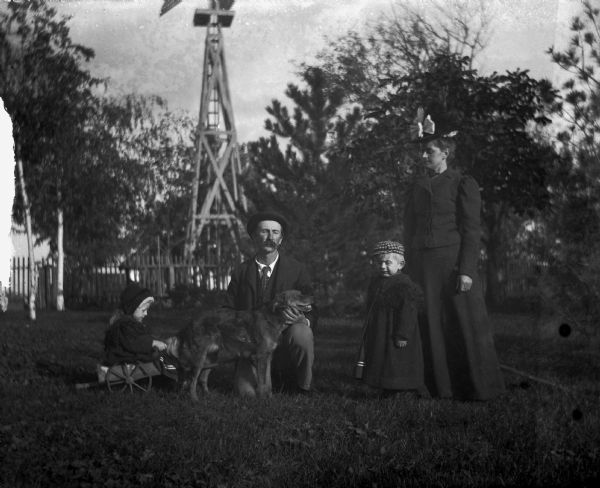 Portrait of the Alex Krueger family posing in yard. From left to right: Jennie, Alex, Edgar, and Florentina Krueger. There is a fence and a windmill in the background.