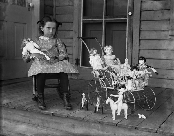 Selma Krueger sitting in a rocker on the porch, holding a doll. Beside her is a buggy filled with dolls and other toys.