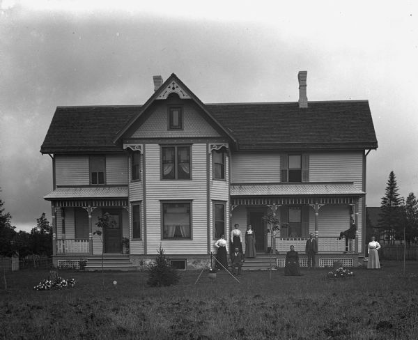 Exterior view of the William Wills homestead. The family, including a dog, is standing around the front porch. One young girl is standing inside the house looking out of the large window on the porch.