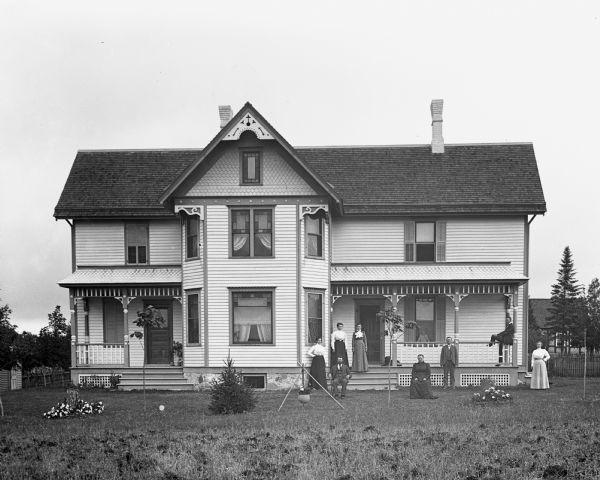 Exterior view of the William Wills homestead. The family, including a dog, is standing around the front porch. One young girl is standing inside the house looking out of the large window on the porch.