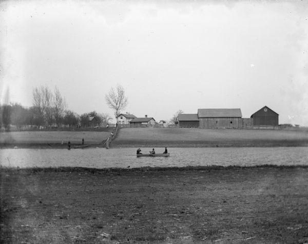 View of the Herman Backhaus farm buildings from across the Rock River. Four people are sitting in a rowboat. Other people are standing along the shoreline and along the fence leading up to the farmhouse.