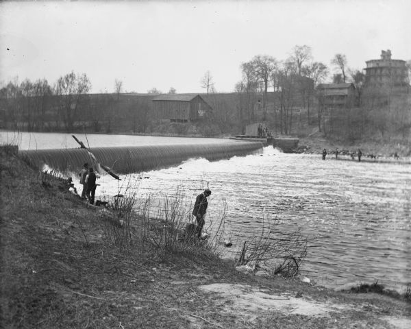 Men standing on either side of the river, fishing near the rough and ready dam. In the background is the Octagon House, built in 1854.