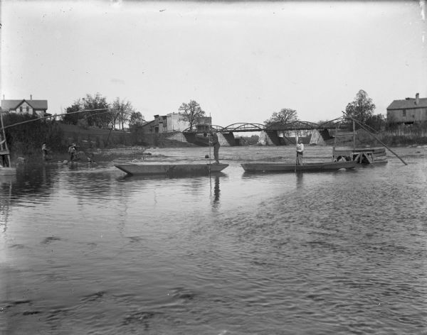 Children are standing along the shoreline watching young boys standing in boats fishing with dip nets in the Crawfish River. In the background is a bridge spanning the river.