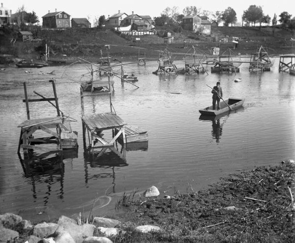 View from shoreline of a man standing in a boat, dipping for carp on the Crawfish River. Behind him dip nets are set up on scaffolds in the river. There are dwellings along a ridge on the far shoreline.
