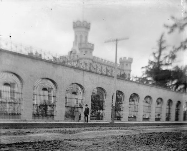 A couple walks outside the gates of the Waupun State Prison, built in 1854. The tower of the central building can be seen beyond the gates.