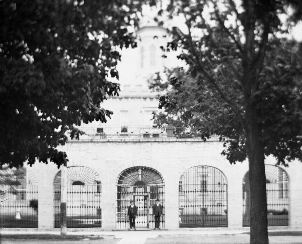 Exterior view of Waupun State Prison, built in 1854. Two men are standing at the main entrance gate. Beyond the prison gates is the tower of the main building.