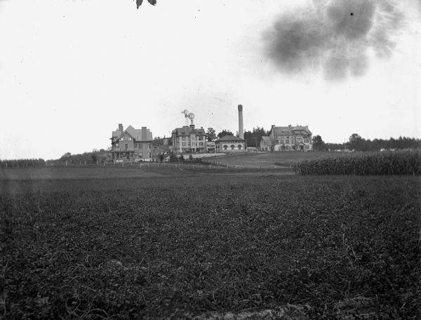 View across fields towards University of Wisconsin Madison buildings. On the left is standing the home of the Dean of the College of Agriculture, Stephen Moulton Babcock (1843-1931), built in 1897. To the right of the Dean's house is the Horticulture Building, with two windmills. Also included are the Wisconsin Dairy School buildings and the heating plant.