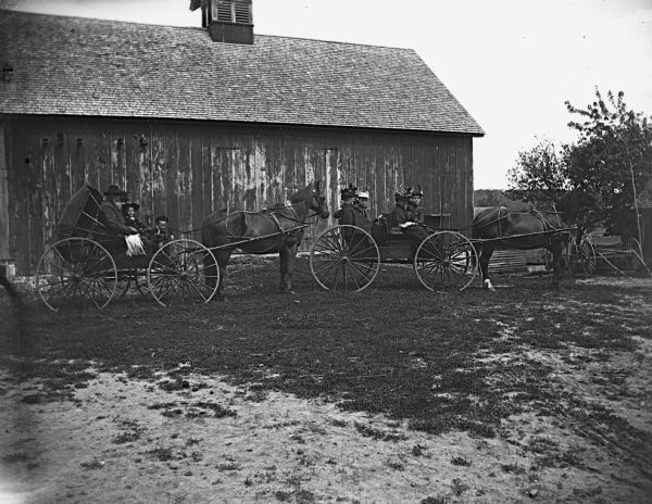 Horse-drawn wagons are lined up in front of an old barn on a Sunday afternoon drive. The wagons carry Mr. and Mrs. August Krueger, and the August Hable family.