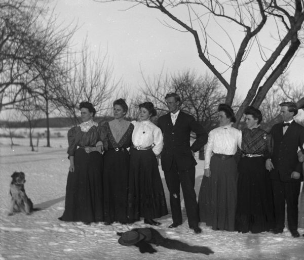 Members of the Krause, Krueger, and Goetsch families standing in the snow with arms linked together. A dog sits to the left, and hats and scarves are lying on the ground in front of the group.