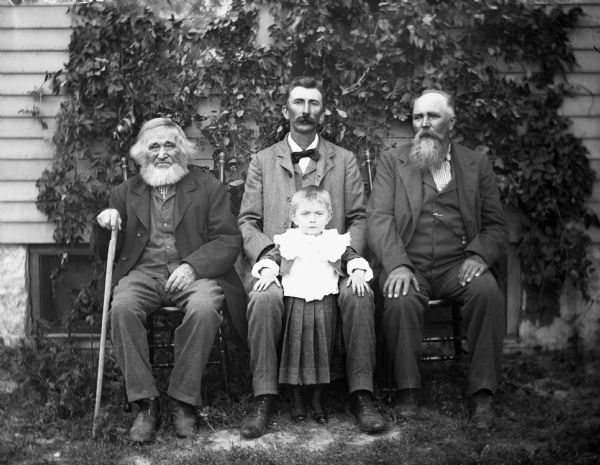Outdoor portrait of four generations of three Krueger men and one boy. The men are sitting in chairs in front of ivy climbing up the side of a house. Sitting from left to right are: William (holding cane), Alexander, and August. Edgar Krueger is standing between his father's legs.