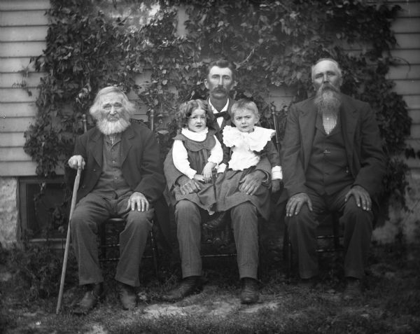 Outdoor portrait of four generations of the Krueger family are sitting in chairs in front of a house with ivy climbing up the side. From left to right sitting in chairs are: William, Alexander, and August Krueger. The twins, Jeannie and Edgar, sitting on Alexander's lap.