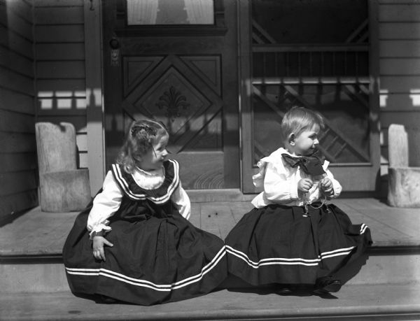 Second birthday portrait of Jennie and Edgar Krueger. The twins are sitting on the top step of a porch, with both wearing dresses. Edgar is holding his fathers watch chain in his hands.
