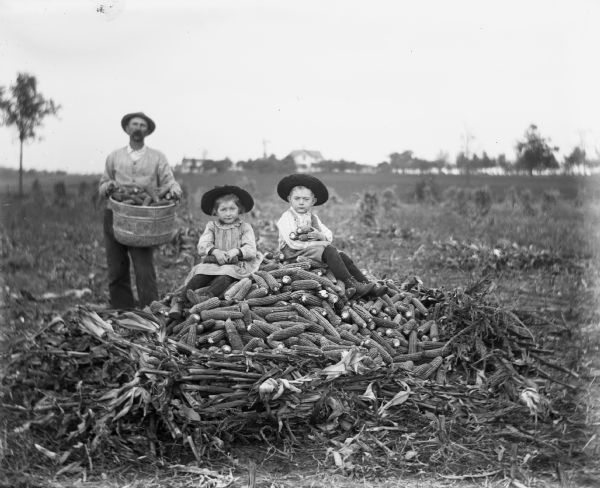 Jennie and Edgar Krueger are sitting atop a pile of husked corn in a cleared field, while Alexander Krueger is holding a pail full of husked corn behind them. The children are each holding several ears of corn in their laps. Corn shocks are scattered throughout the cleared field.