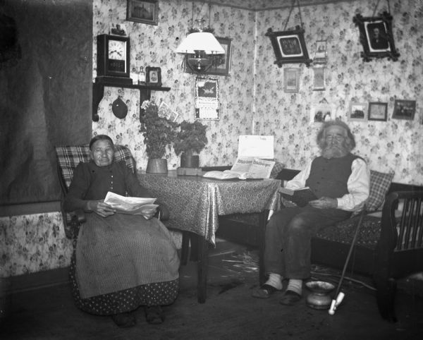 Indoor flash portrait of William and Johanna Krueger sitting in the living room. Johanna is reading the newspaper, while William is reading a book. Various family portraits are hanging along the walls. A cane and a spittoon are resting on the floor next to William.