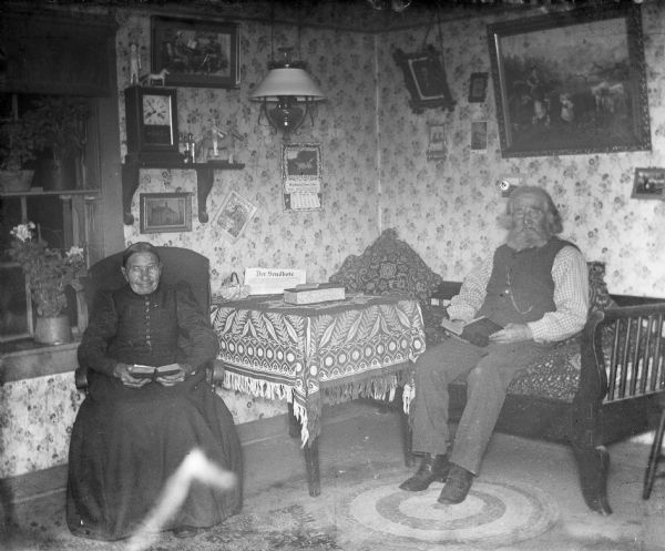 Indoor flash portrait of William and Johanna Krueger sitting in the living room reading books. Various calendars, figurines, and portraits are hanging along the walls.