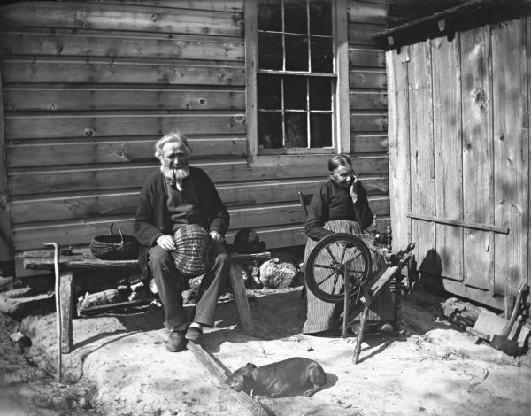 William and Johanna Krueger are sitting alongside a house near a shed. William is sitting on a bench weaving a willow basket, and has a pipe in his mouth. Johanna is sitting in a chair next to him spinning yarn with a spinning wheel. A small dog is laying on the ground in front of them.