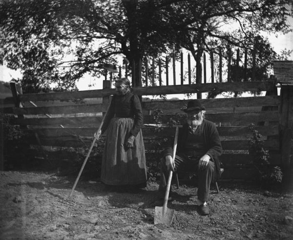 William and Johanna Krueger working in the garden. Johanna is standing next to the garden fence holding a hoe. William is sitting next to her on a small bench, smoking a pipe and holding a shovel.