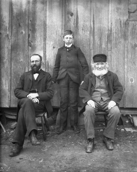 Outdoor portrait of three generations of Krueger men standing next to a house. Albert and William are sitting in chairs. Ben Krueger is standing between his father and grandfather.