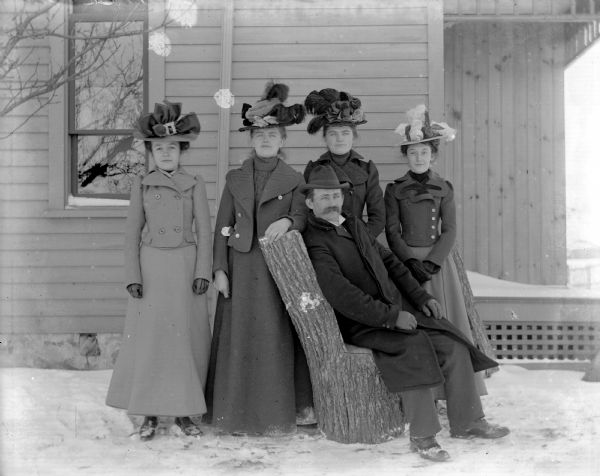 Outdoor winter group portrait of Herman Reibe and his four sisters.  Herman is sitting on a log that has been fashioned into a chair, while his four sisters are standing behind him wearing elaborate hats.