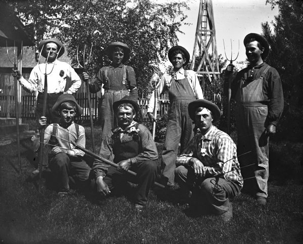 Group of seven men, each holding a pitchfork, standing in a yard.  Front row, left to right: Herman Bentert, Herman Riebe, Henry Burchardt. Back row, left to right: Willie Will, Willie Wendorf, Henry Reusak, and Clancy Fairfield. In the background is a wooden tower structure, perhaps for a windmill.