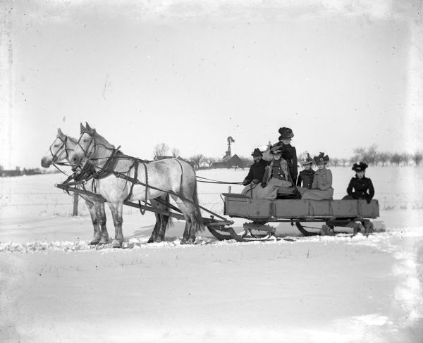 Winter scene with Herman Riebe, his four sisters, and a cousin riding in a bobsled pulled by a team of white horses through the snow. A fence, a snowy field, farm buildings and a windmill are in the background.