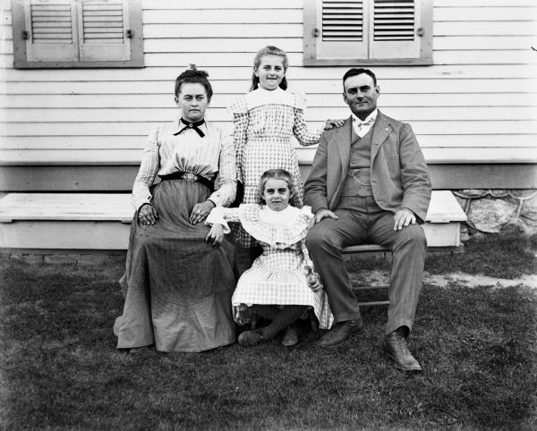 Outdoor family portrait, in the yard of a house, of the Fels family. George and his wife, Ida, are sitting in chairs, while their two daughters, Flora and Hattie, are situated between them.