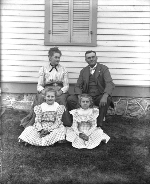 Outdoor family portrait of the Fels family. George and Ida Fels are sitting in chairs next to the side of a house, while their two daughters Flora and Hattie are sitting on the ground in front of them.