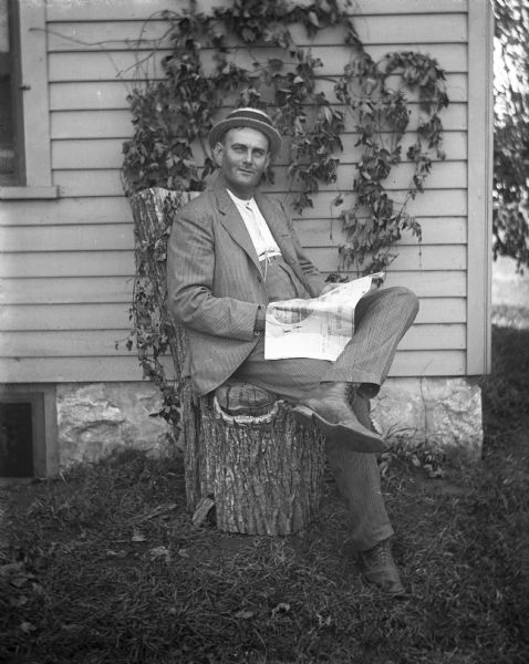 Outdoor portrait of George Fels sitting on a chair reading a newspaper near the corner of a house. Ivy is growing up the side of the house, and the chair George is sitting on is made from a log.