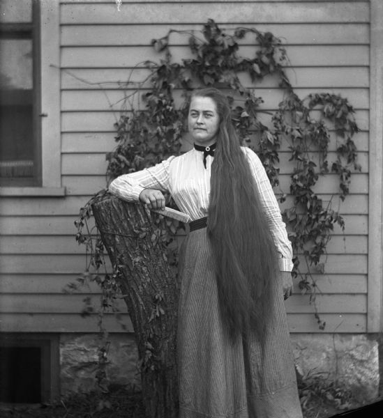 Outdoor portrait of Ida Fels leaning against a log chair with her long hair draped over her left shoulder. In her right hand she is holding a comb. She is standing in front of the side of a house with ivy crawling up the siding.