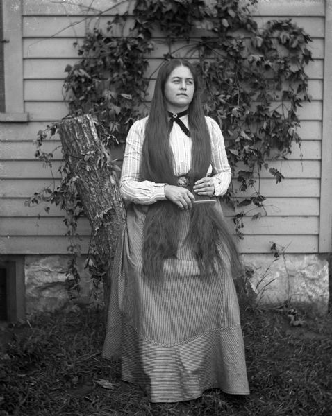 Outdoor portrait of Ida Fels leaning against a log placed next to the side of a house with ivy crawling up the side. She is combing her long hair, which is hanging down over her shoulders.