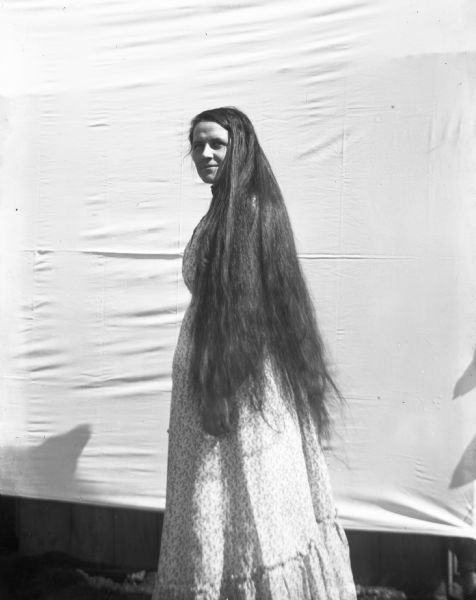 Ida Fels (?) standing in front of a white backdrop looking over her shoulder at the camera with her long hair let down.