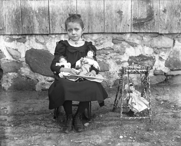 Hattie Pautz, sitting and posing with her dolls in front of the side of a building. On the ground next to her is one of the dolls in a doll-sized lawn swing.