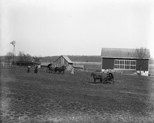 View across pasture towards two horse-drawn carriages on the August Pautz farm. Two people are sitting in each carriage, and two women and a child are standing on the left. A farm house and farm buildings are in the background.
