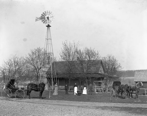 Members of the Pautz family lined up in front of their house. Two carriages with horses are flanking either side of the family. A windmill is situated alongside the house.