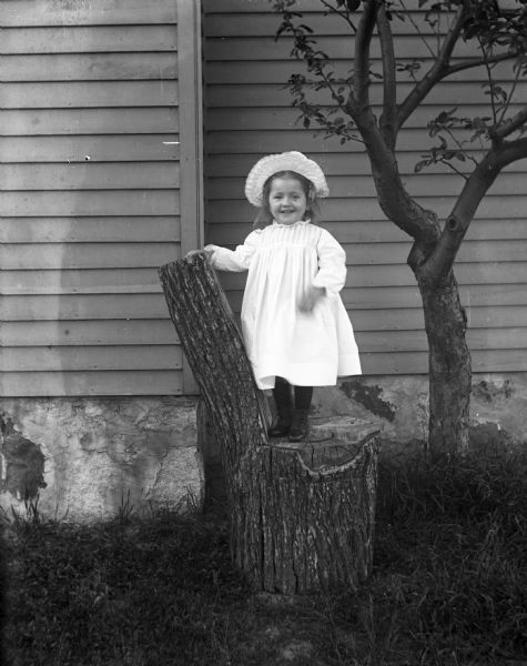 Jennie Krueger standing on a chair made from a stump in the yard in front of the side of a house.
