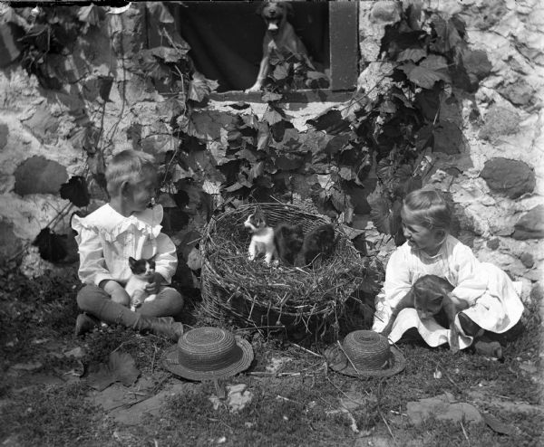 Jennie and Edgar Kueger are sitting cross-legged next to a house with their hats on the ground in front of them. They are both smiling and looking at a basket between them that is filled with hay and three kittens. A kitten is sitting in Edgar's lap, while the mother cat is sitting in Jennie's. A dog is standing and looking out of a window above the basket.