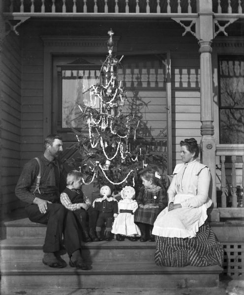 The Alexander Krueger family sitting on a porch in front of a decorated Christmas tree. Alexander and Edgar are sitting on the left side of the porch steps, while Florentina and Jennie are sitting on the right. Two dolls are sitting between the twins.