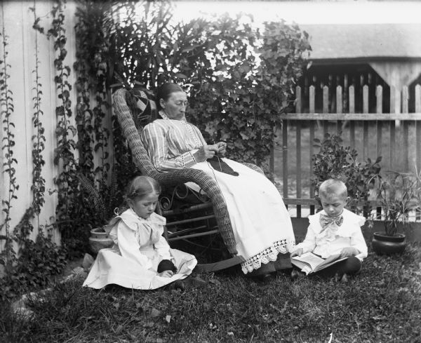 Mary Krueger knitting in a rocking chair in the yard while her grandchildren are sitting on the ground beside her. Jennie Krueger is sitting to her right, holding the remaining yarn. Her brother, Edgar, is sitting to the left of Mary reading a book.