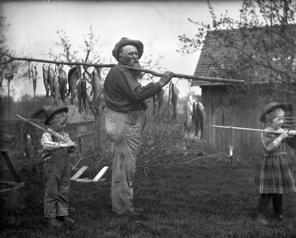 Jennie and Edgar Krueger are with their grandfather, August Krueger, who is carrying fresh fish. The two children each have one fish attached to a pole they are holding over their left shoulder. August is carrying a bigger pole over his left shoulder with twenty fish attached.