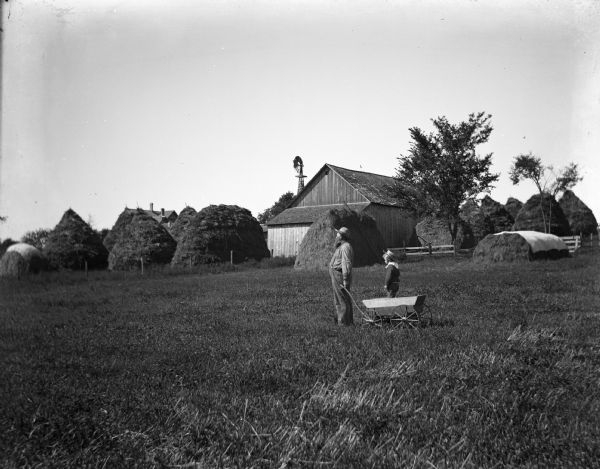 August and Edgar Krueger pulling a small wagon through an open field. A barn and large haystacks run along the perimeter of the field.