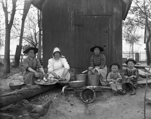 Florentina, Sarah, Mary, Edgar, and Jennie Krueger are sitting in front of a small building cleaning fish. Florentina and Sarah are scaling fish, while Mary is holding a knife and gutting the fish. Edgar and Jennie are each holding a fish in their laps. A chicken is standing nearby.