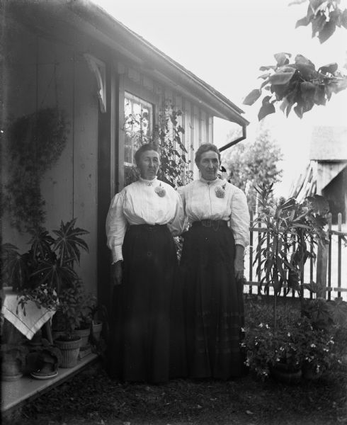 Mary Krueger standing with her sister Emil Bigalk amongst potted plants near the side of a house. They are each wearing an identical corsage on their left chest.