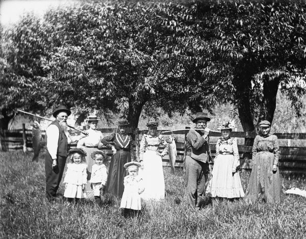August, Florentina, Mary, Jennie, and Edgar Krueger with members of the Ulirich family standing near a fence, returning from spear fishing for carp. August and Mr. Ulrich are each carrying a carp hanging from a stick over their shoulder.