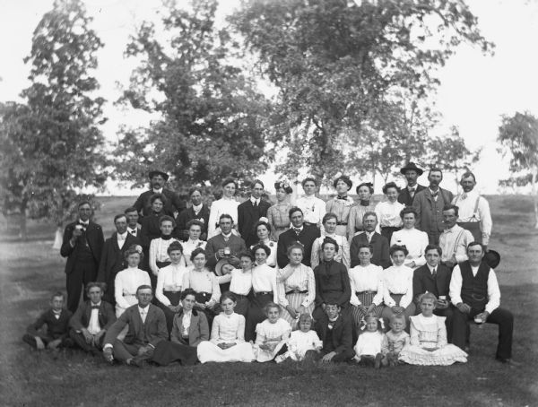 Large group portrait of members of the community attending a picnic at the Wills family woods.  Hattie Fels, Edgar Krueger, and Jennie Krueger sit in the front row on the right, while Flora Fels sits sixth from the right.  In the second row George Fels sits on the right end, while Florentina Krueger sits fourth and Ida Fels sits sixth from the right. Alexander Krueger stands second from right in the back row.