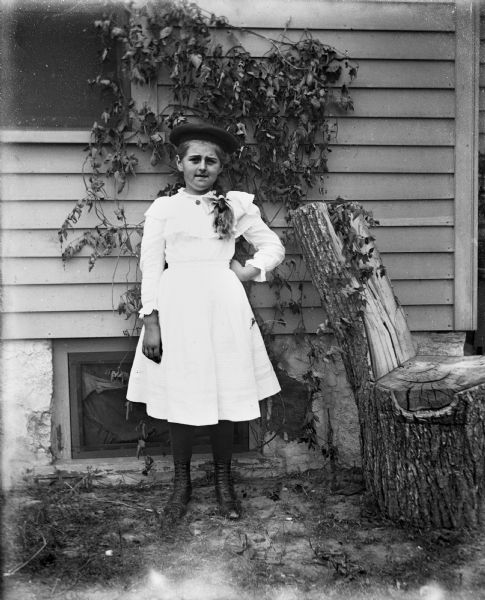 Outdoor portrait of Hattie Fels standing next to a chair made from a log stump, which is placed close to the side of a house with ivy climbing up the side.