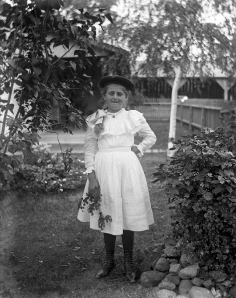Outdoor portrait of Hattie Fels standing in a yard between a small tree and a shrub. She is holding a branch in her right hand.